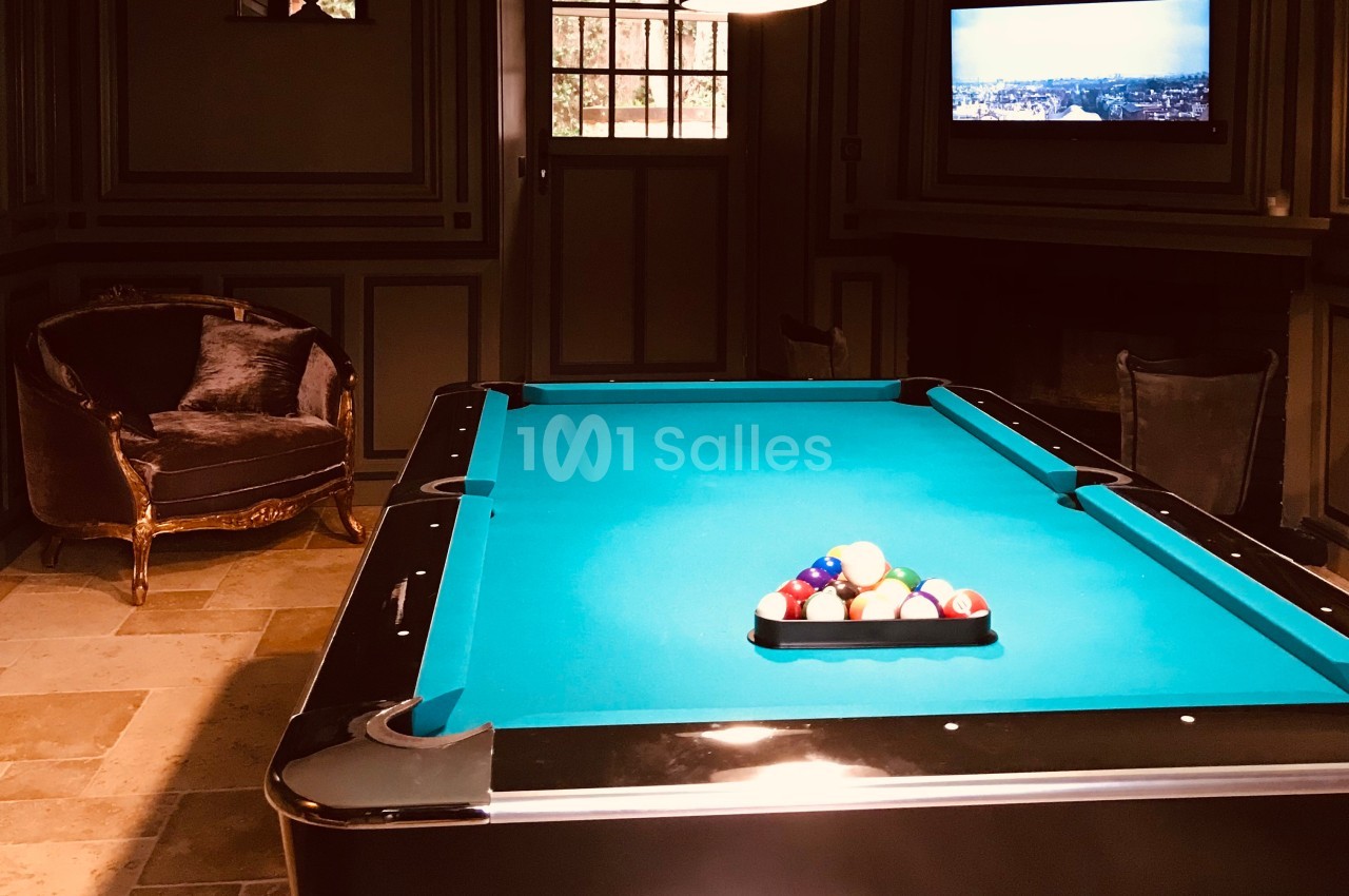 Table de billard avec des boules alignées, située dans une pièce sombre avec fauteuil, fenêtre et télévision murale.