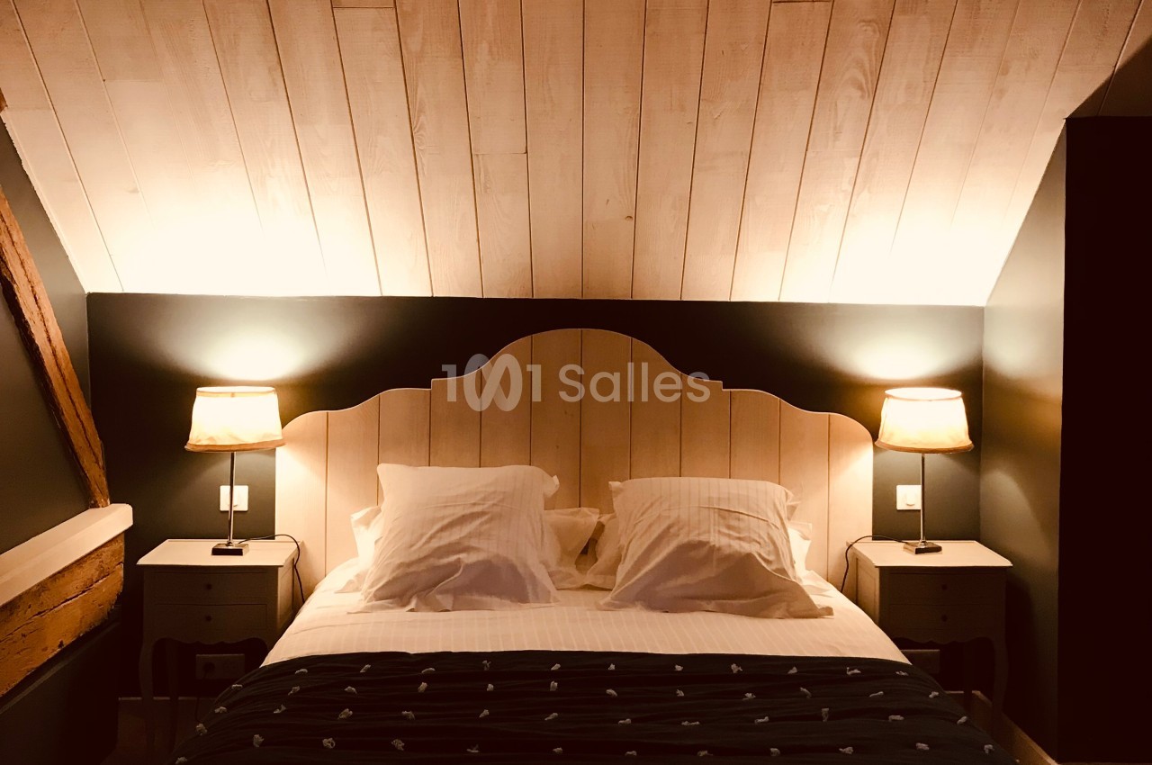 Chambre avec lit double, draps blancs, plaid sombre, deux lampes de chevet allumées et plafond en bois clair.