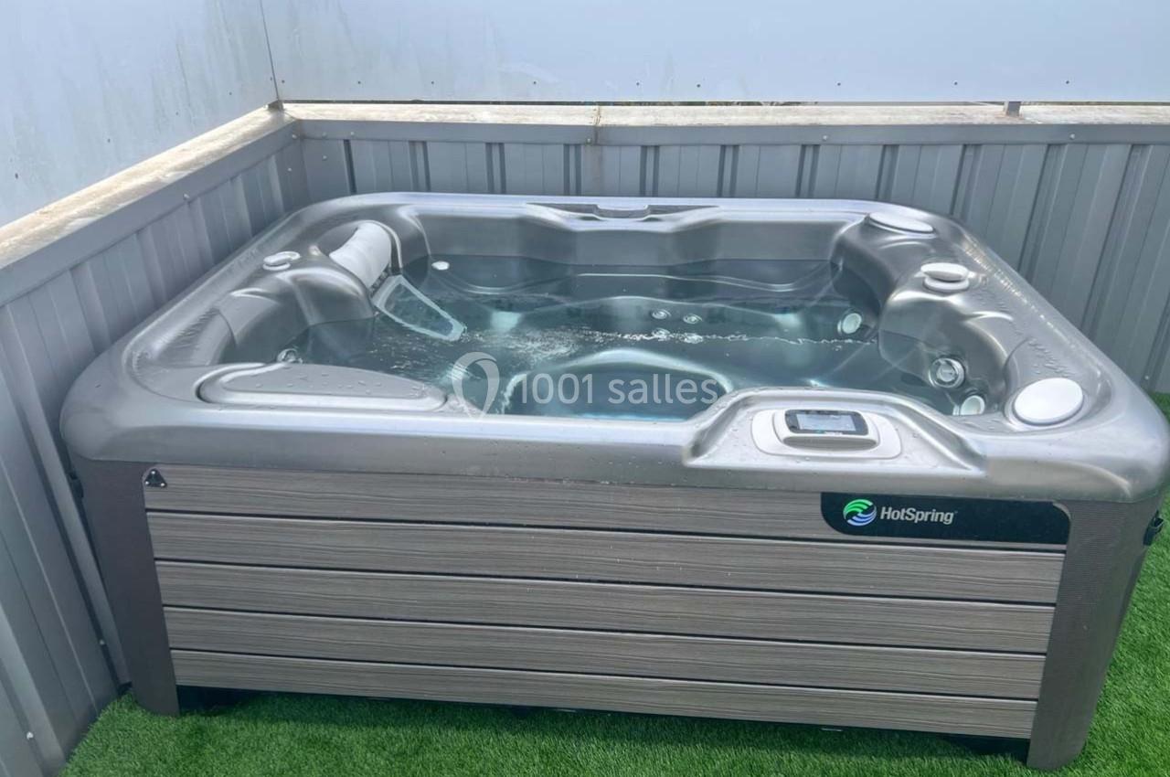 Spa extérieur gris rempli d'eau, installé sur une pelouse artificielle et entouré de panneaux métalliques.