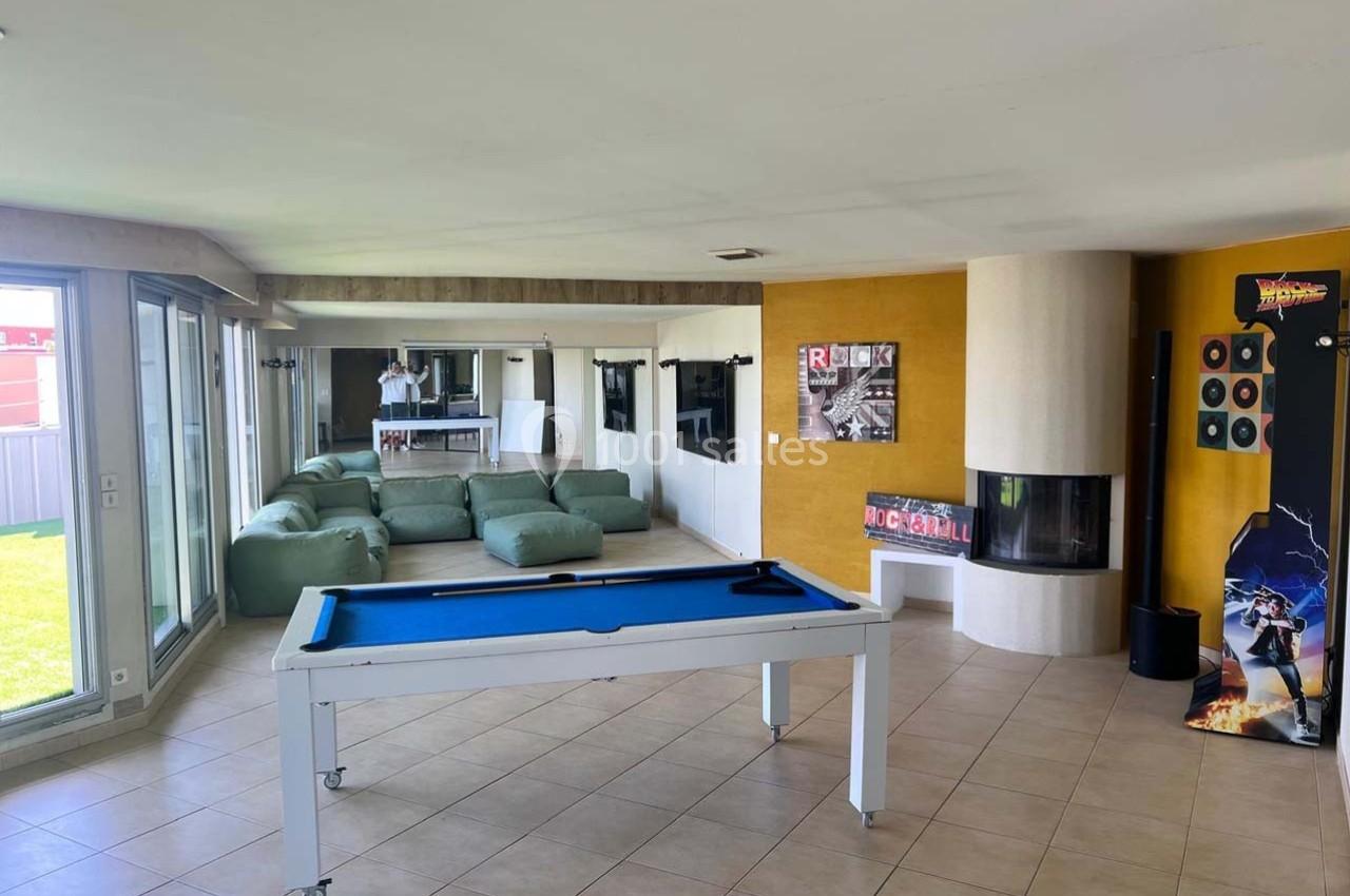 Salle de loisirs lumineuse avec billard, poufs verts, cheminée moderne et décorations murales.