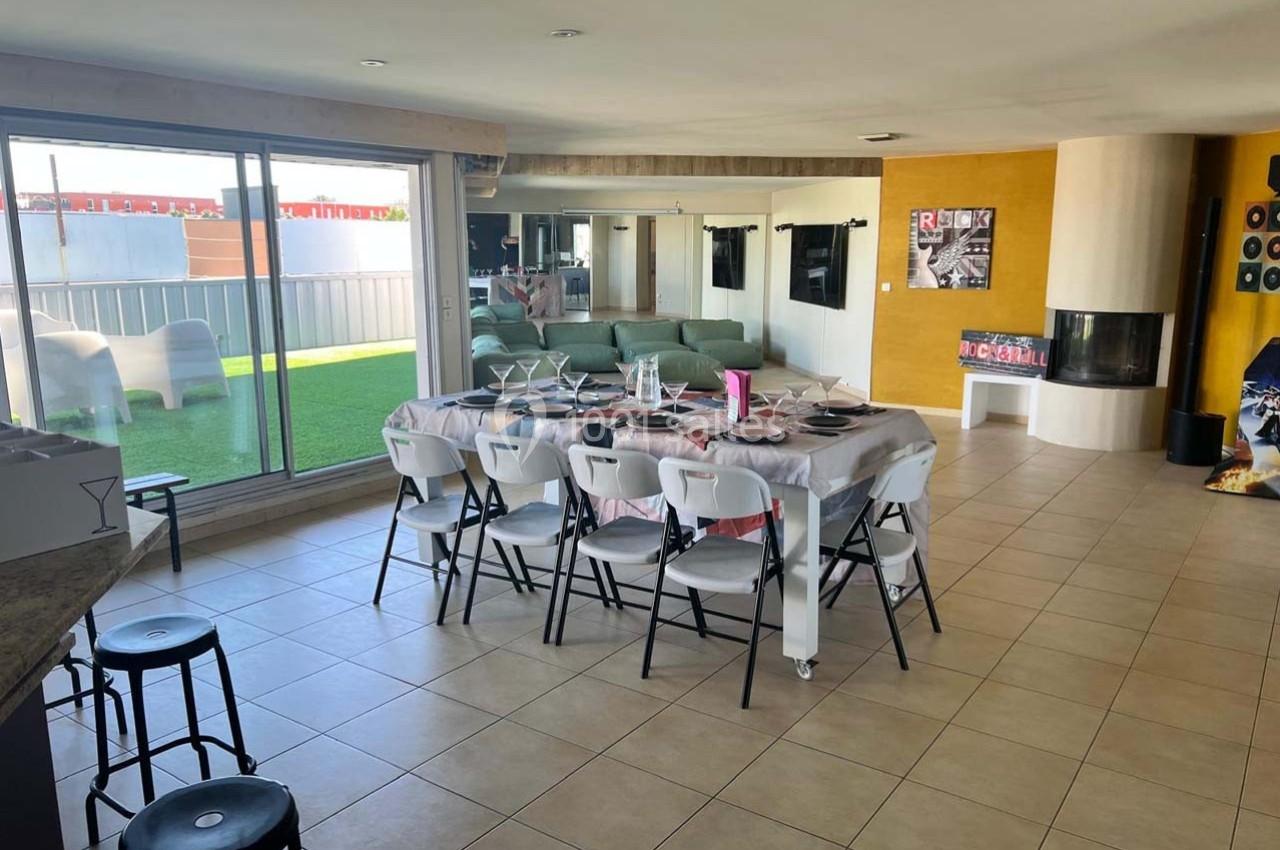Salle à manger lumineuse avec une grande table dressée, un salon en arrière-plan et une terrasse extérieure visible.