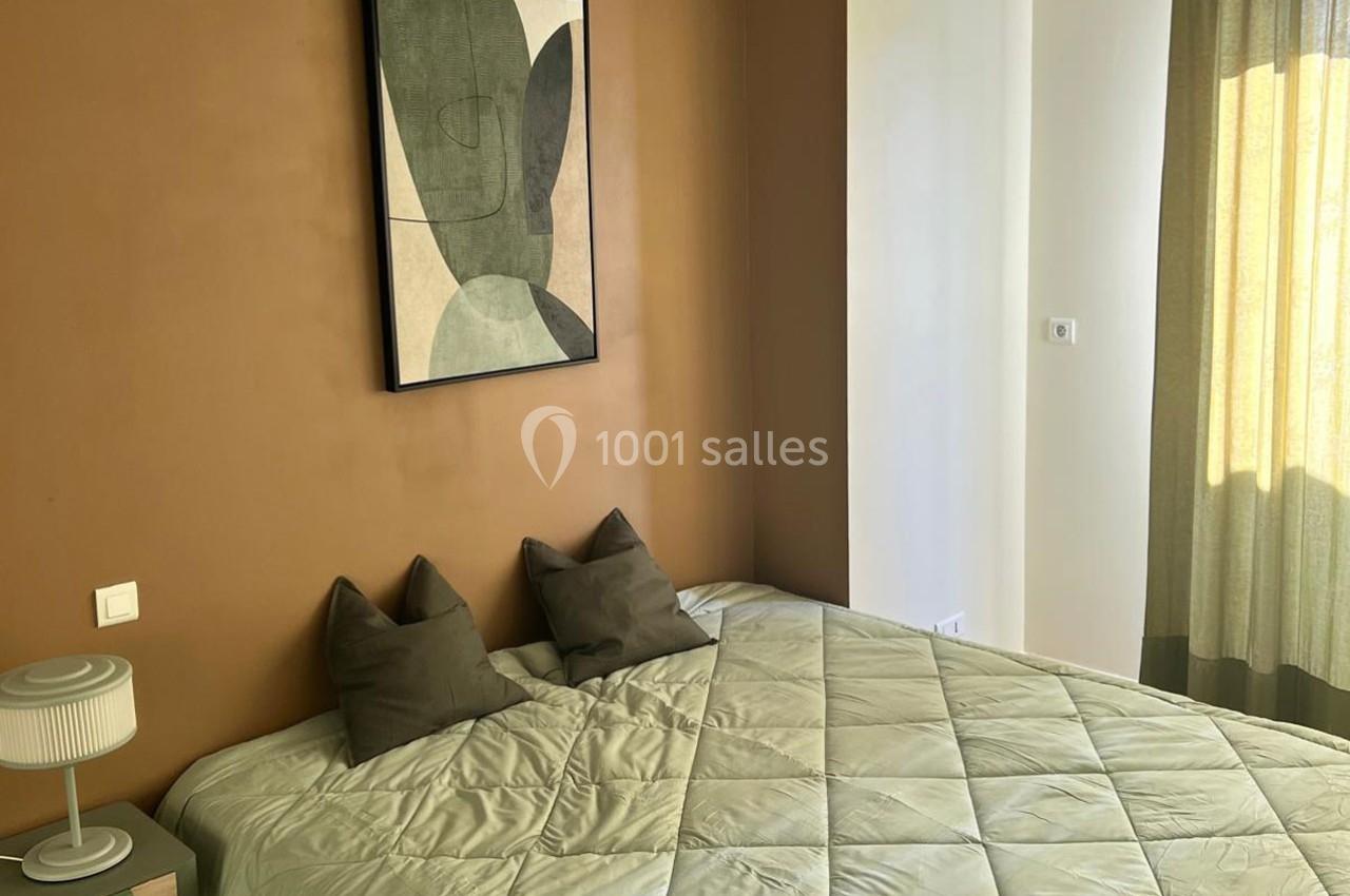 Lit double avec couverture beige, deux coussins gris, lampe de chevet et tableau abstrait sur un mur marron.