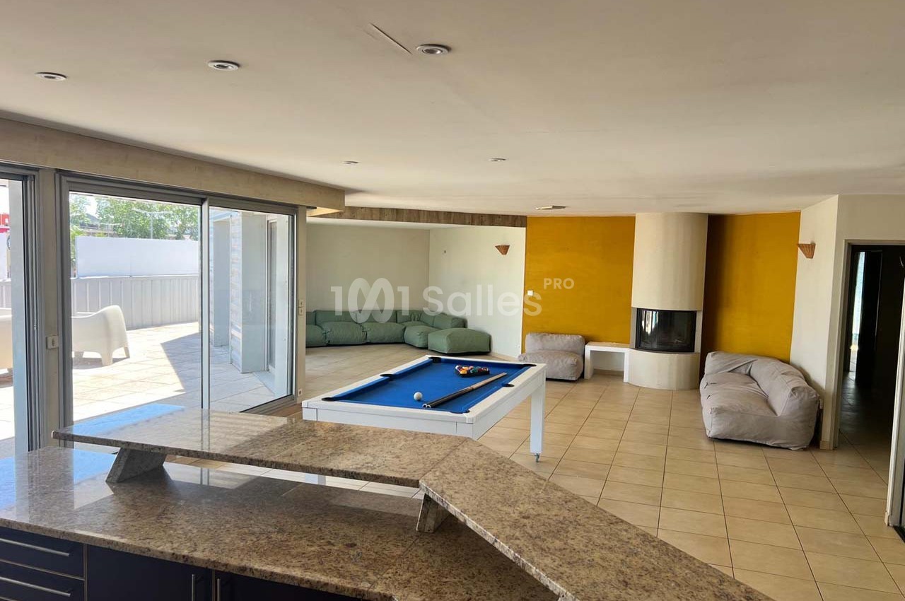 Salle lumineuse avec billard, canapé d'angle, cheminée moderne, baies vitrées et comptoir en pierre.
