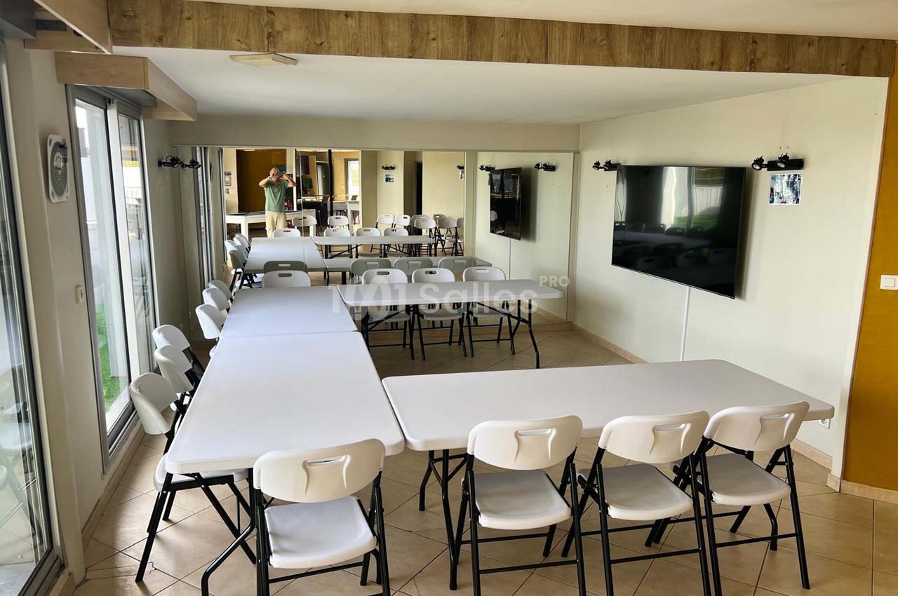 Salle lumineuse avec tables blanches disposées en U, chaises pliantes, grand écran mural et miroirs réfléchissants.