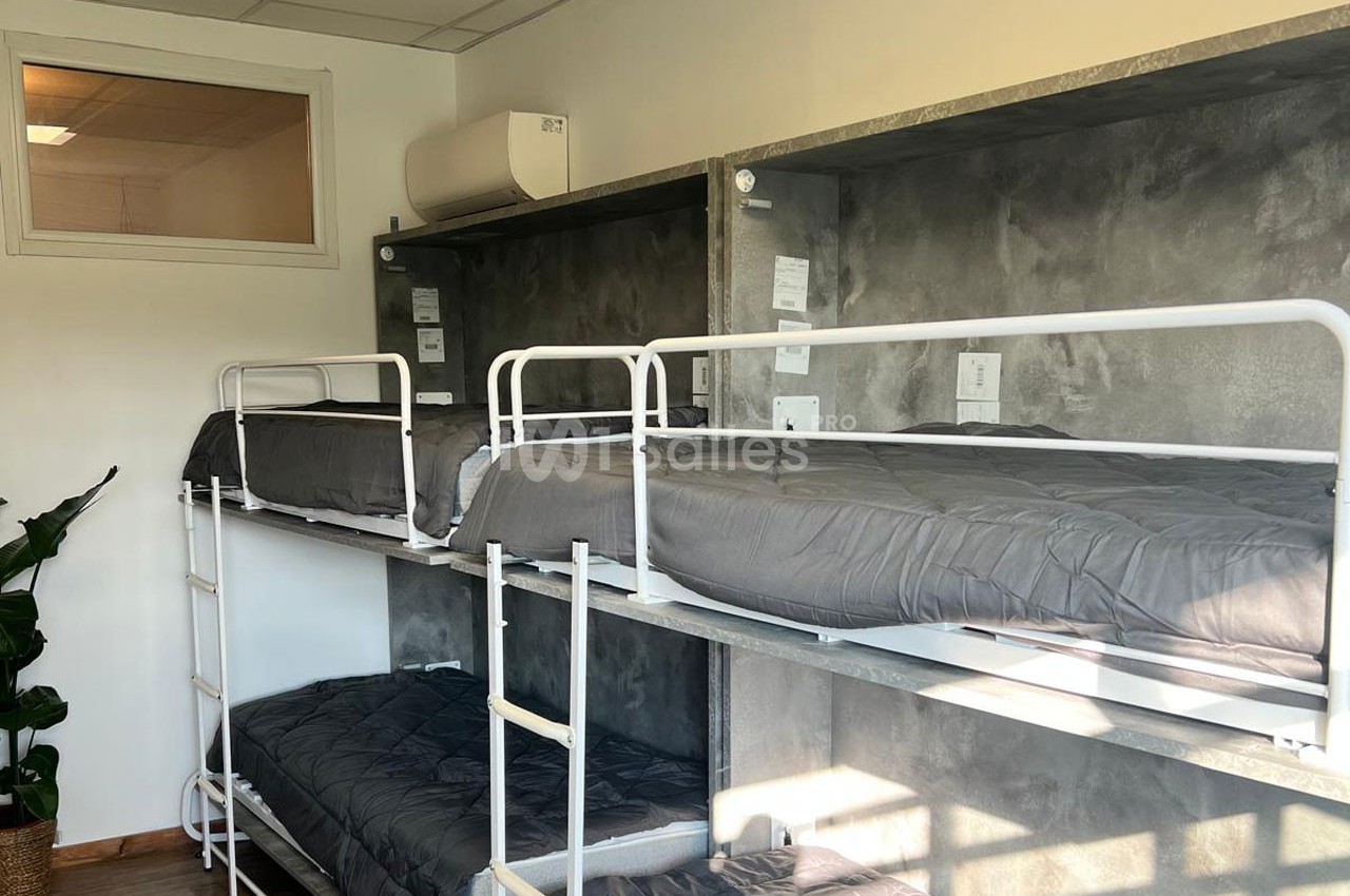 Dortoir avec lits superposés en métal, matelas gris et mur en béton, éclairé par une fenêtre intérieure.