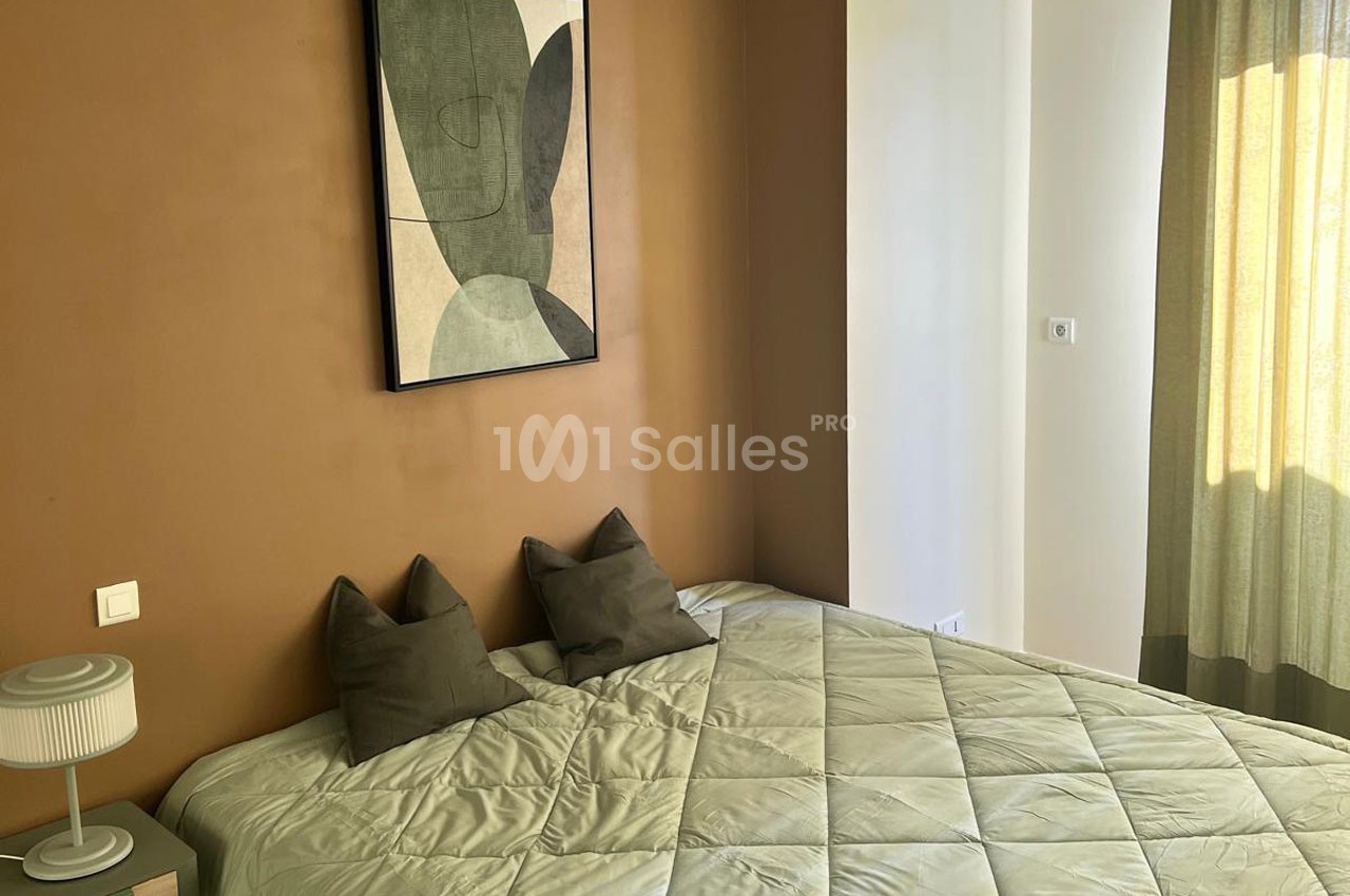 Lit double avec coussins sombres, lampe de chevet et tableau abstrait sur un mur marron, près d'une fenêtre avec rideaux.