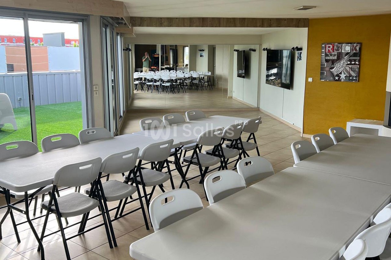 Salle lumineuse avec de grandes tables et des chaises pliantes blanches, donnant sur un espace extérieur avec pelouse.