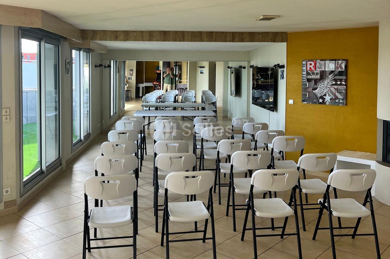 Salle lumineuse avec des chaises blanches alignées en rangées, face à un espace aménagé avec des tables et des affiches au…