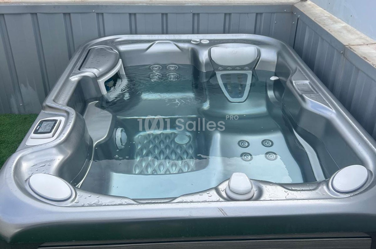 Spa extérieur gris rempli d'eau, installé sur une surface en gazon synthétique et entouré de parois métalliques.