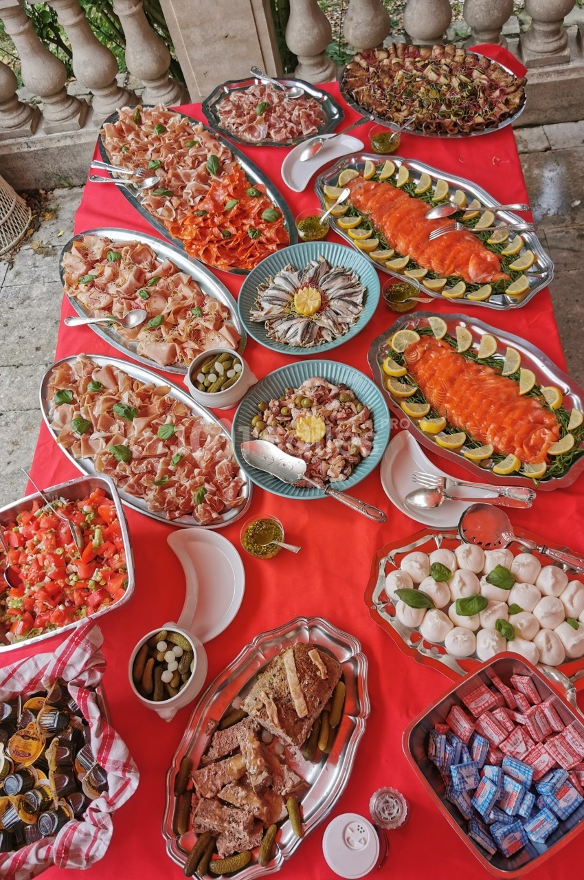 Buffet varié avec charcuterie, fruits de mer, légumes, sauces et pains disposés sur une nappe rouge en extérieur.