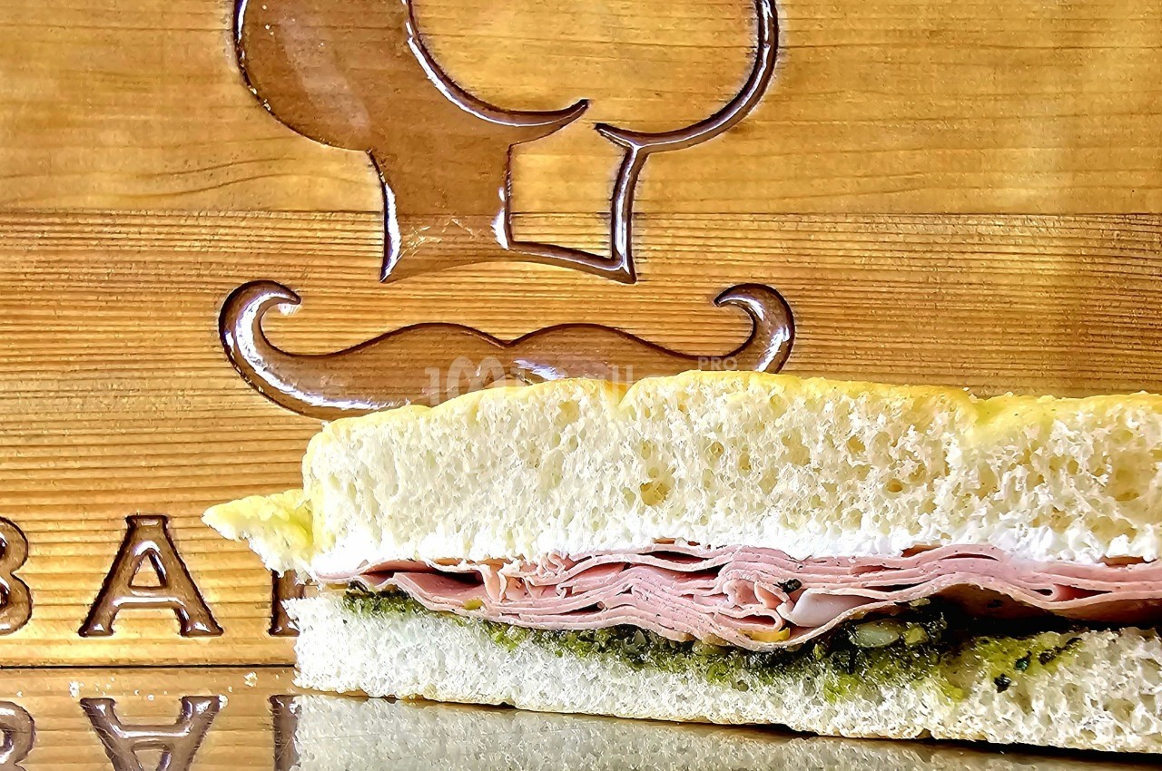 Sandwich garni de jambon et de pesto, présenté devant une planche en bois gravée d'un motif de chef.