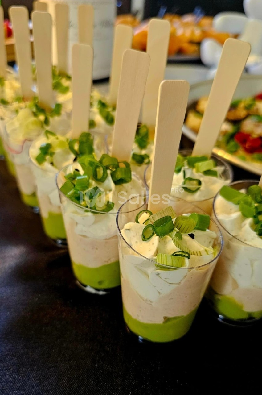 Verrines garnies de mousse verte, crème blanche, ciboulette et bâtonnets en bois, présentées sur une table.
