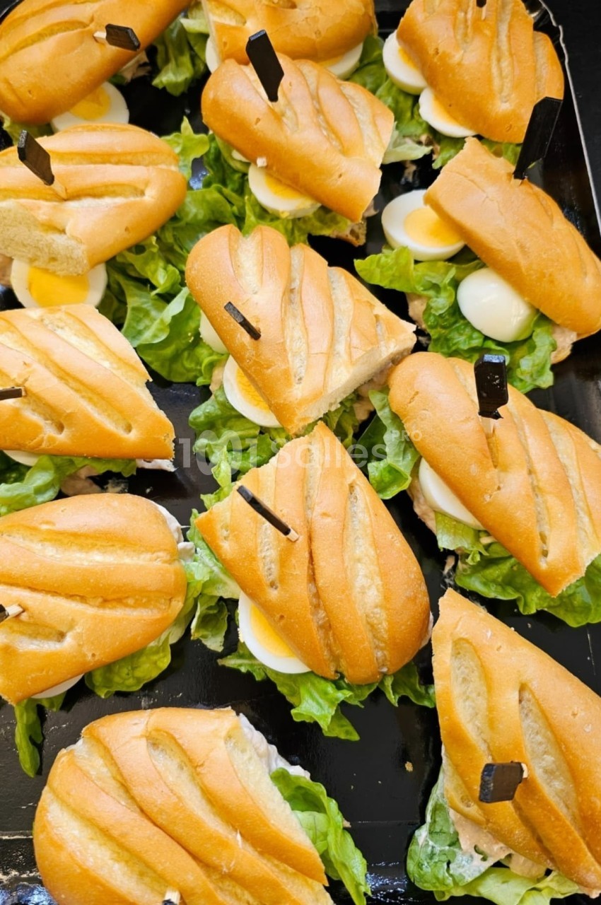 Plateau de mini-sandwichs garnis de salade, œufs durs et sauce, présentés sur un fond noir.