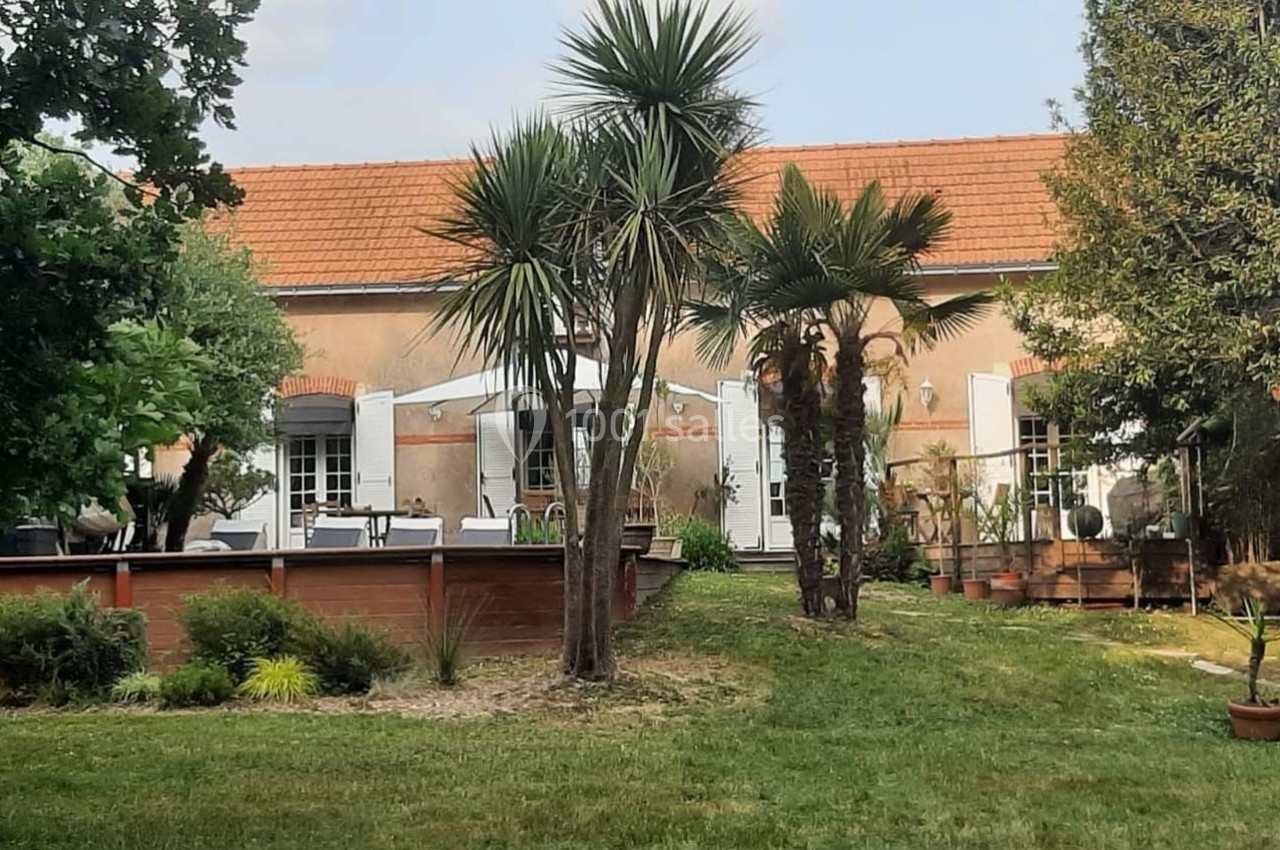 Façade d'une maison avec terrasse en bois, entourée de palmiers et d'arbustes, dans un jardin verdoyant.