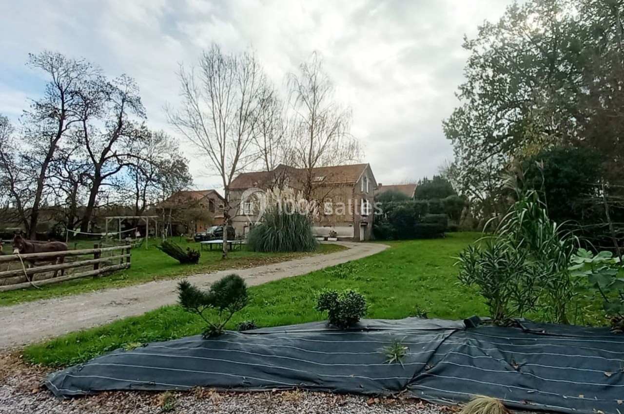 Maison en pierre entourée de verdure, avec un chemin de gravier, des arbres et un jardin en premier plan.