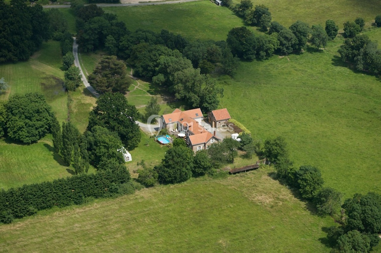 Vue aérienne d'une maison avec toit en tuiles, piscine et dépendances, entourée de champs et d'arbres.