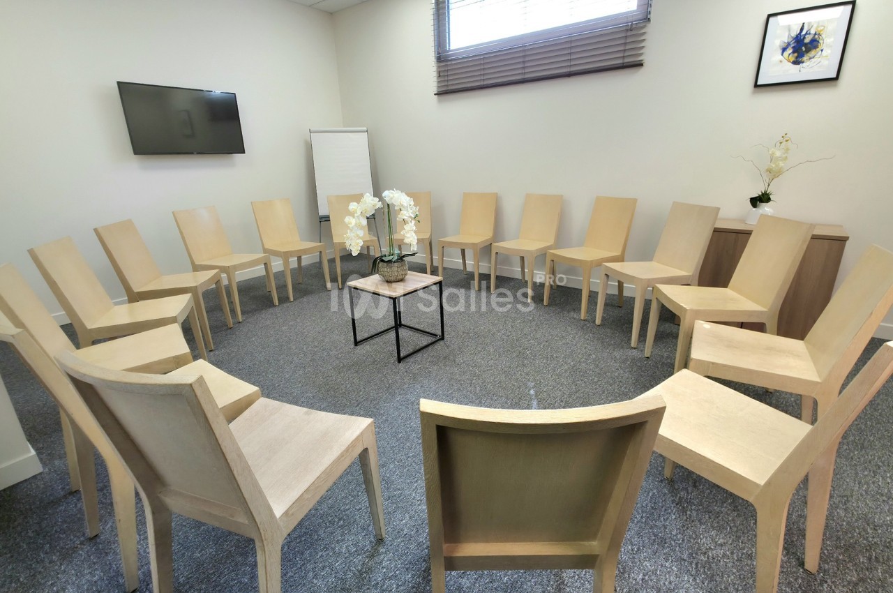 Salle de réunion avec des chaises disposées en cercle, une table basse au centre et un écran mural.