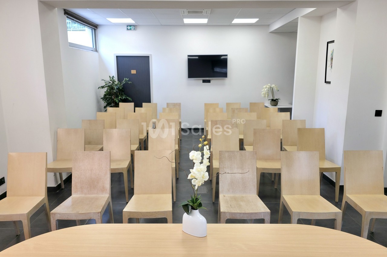 Salle de réunion avec des chaises en bois alignées, un écran mural et une table avec une orchidée au premier plan.