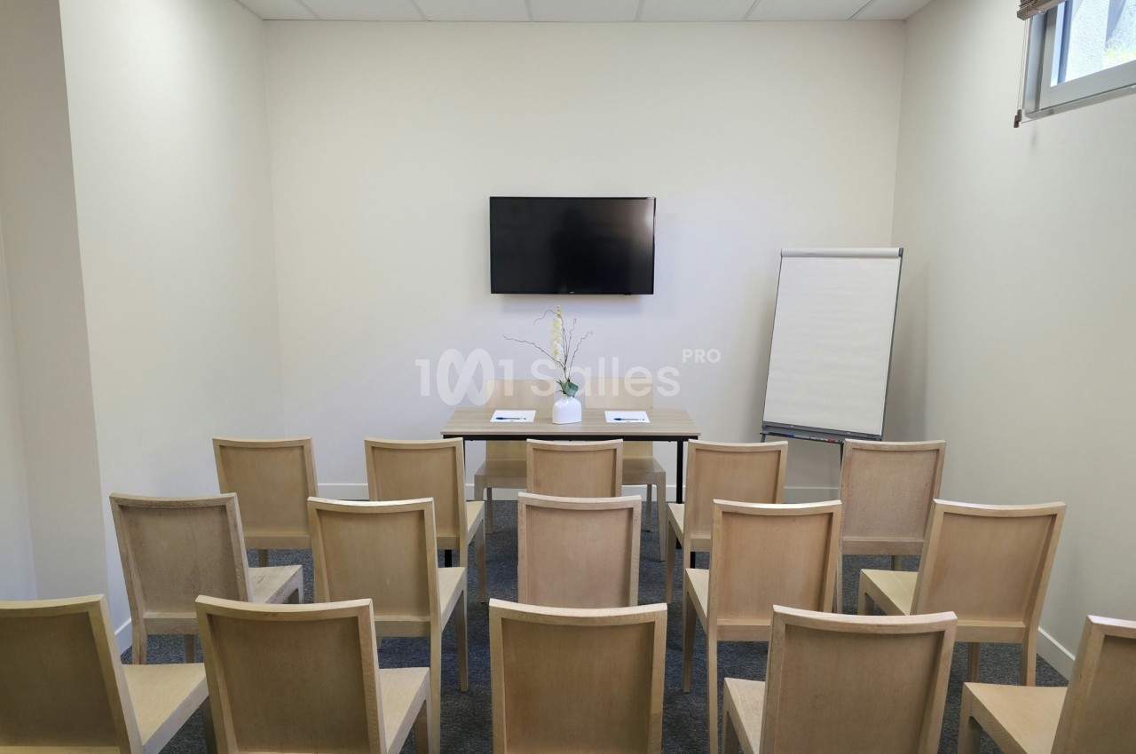 Salle de réunion avec chaises alignées, table, écran mural, paperboard et vase contenant des branches décoratives.