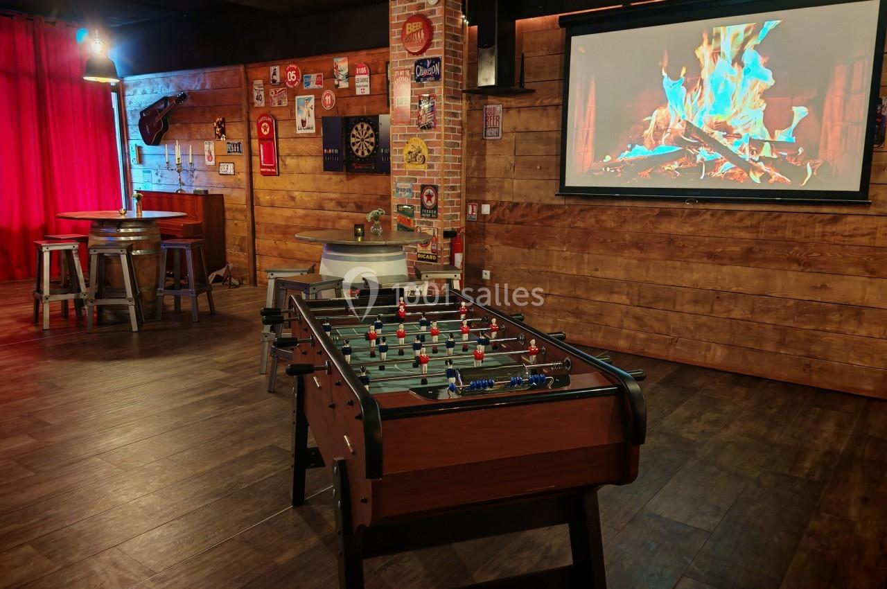 Salle de loisirs avec baby-foot, écran affichant un feu de cheminée, murs en bois et décorations rétro.