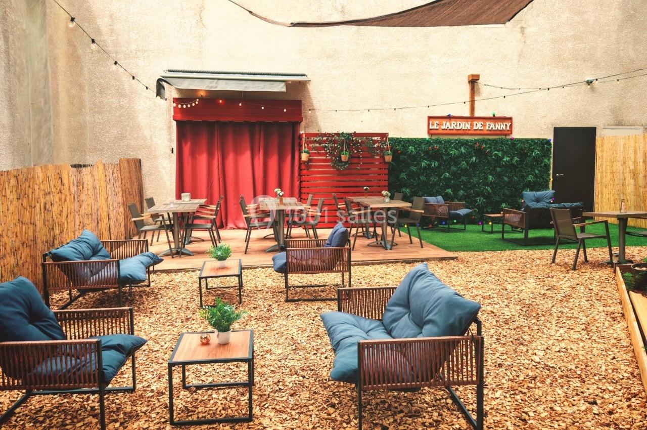 Terrasse extérieure avec chaises et tables en bois, sol recouvert de copeaux, décor végétal et enseigne rouge au fond.
