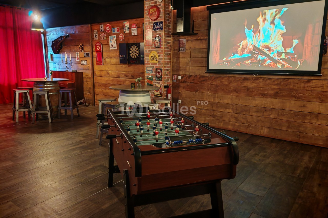 Salle de loisirs avec baby-foot, écran affichant un feu de cheminée, murs en bois et décorations rétro.
