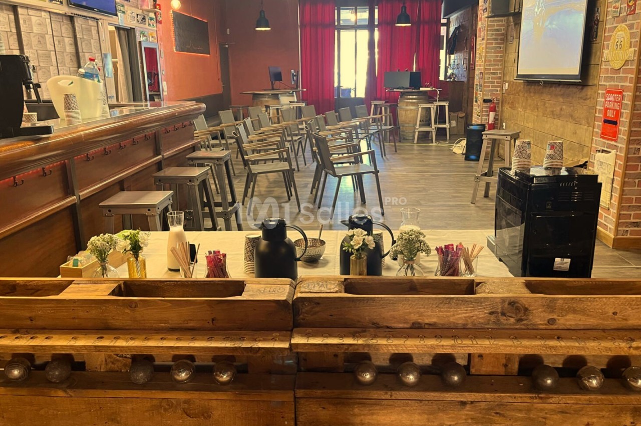 Salle aménagée avec des chaises disposées face à un écran, un bar en bois au premier plan avec des boissons et des…