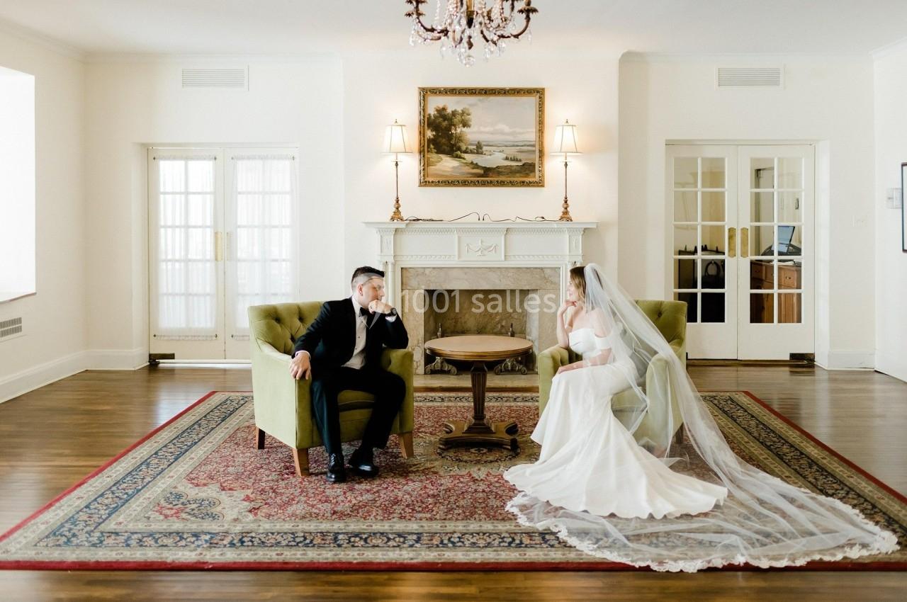 Un couple en tenue de mariage assis face à face dans un salon élégant avec cheminée et lustre.