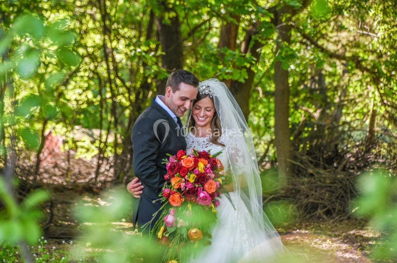 Un couple en tenue de mariage pose dans un jardin verdoyant avec un grand bouquet de fleurs colorées.