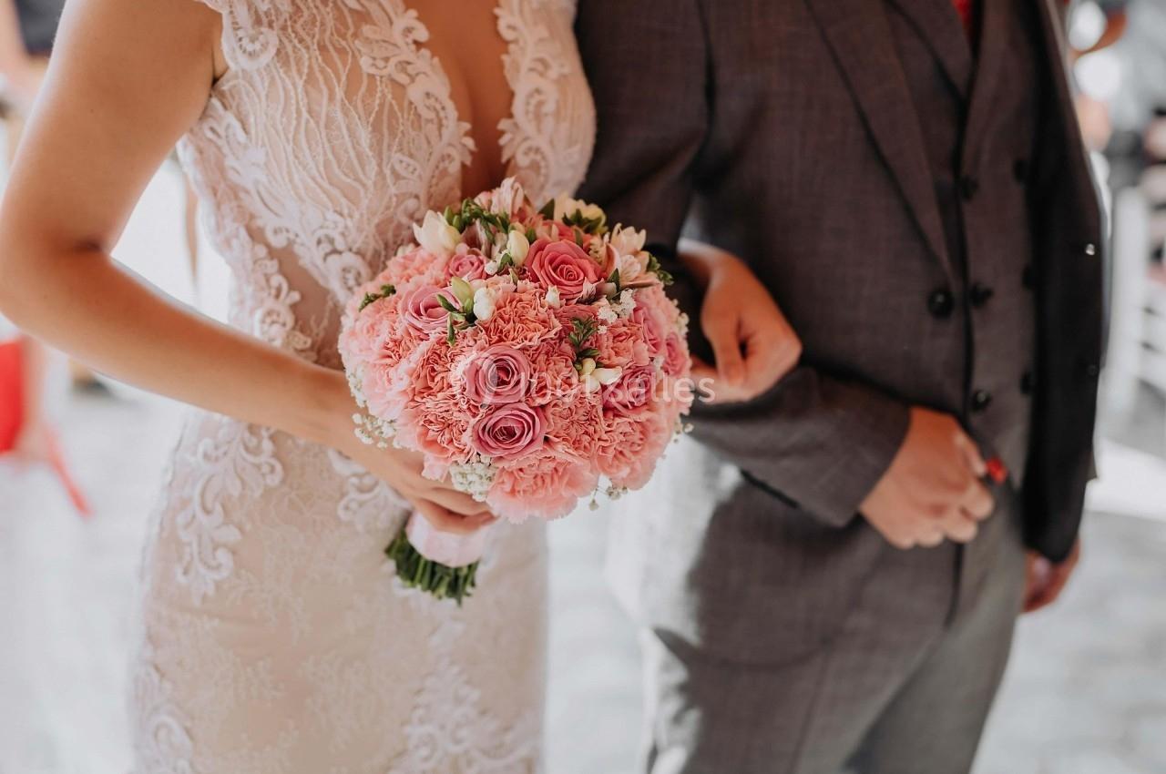 Une mariée en robe blanche tient un bouquet de fleurs roses, accompagnée d'une personne en costume gris.