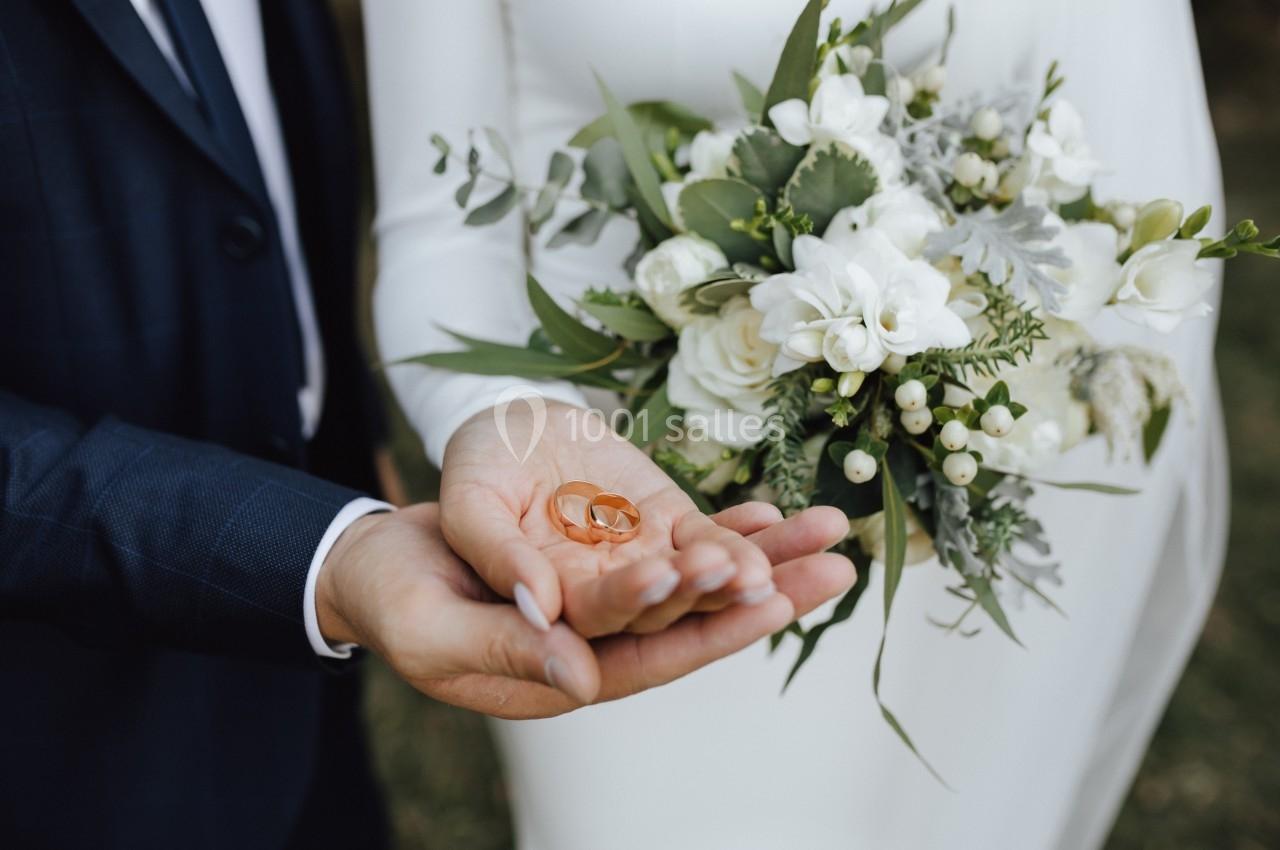 Deux alliances dorées posées dans la main d'un homme en costume, avec un bouquet de mariée en arrière-plan.