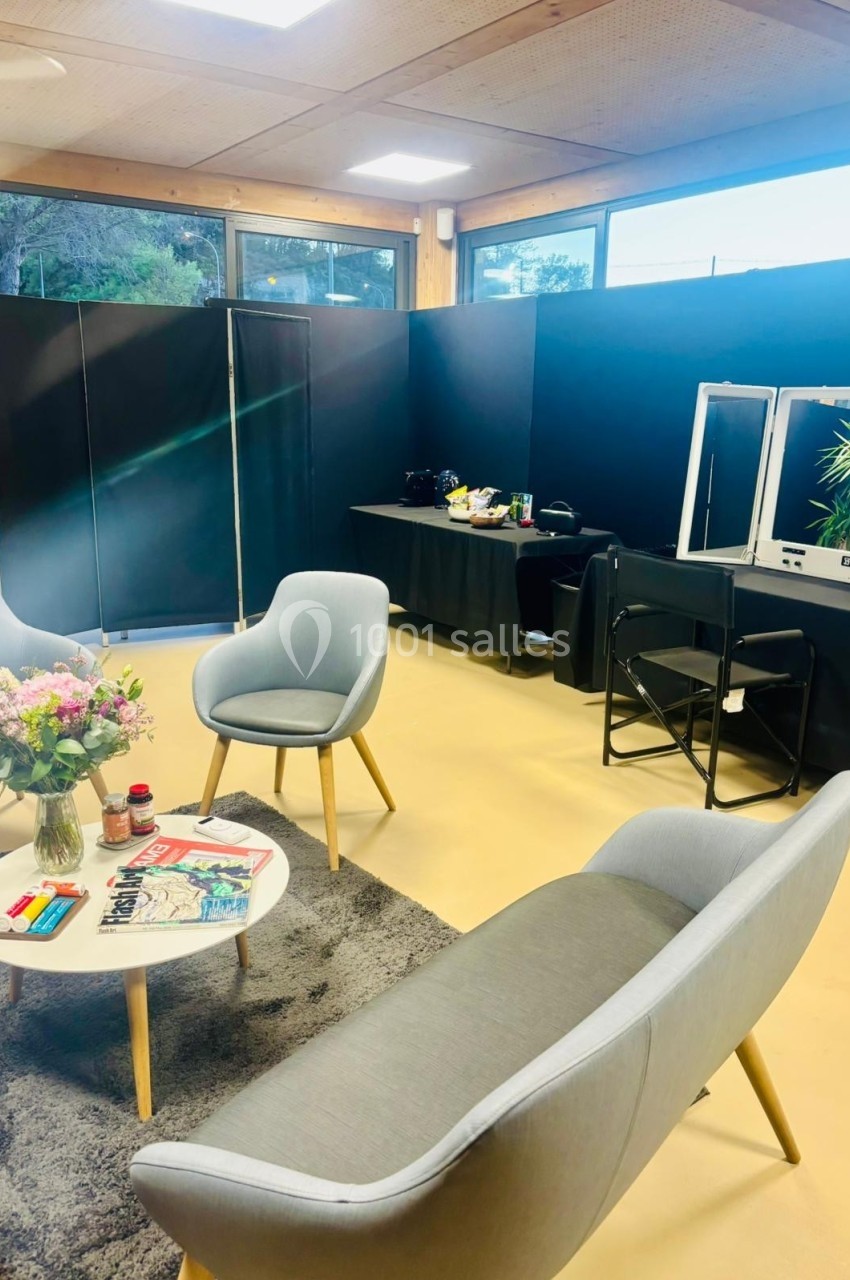 Espace aménagé avec fauteuils, table basse, fleurs, magazines et coiffeuse dans une pièce lumineuse.