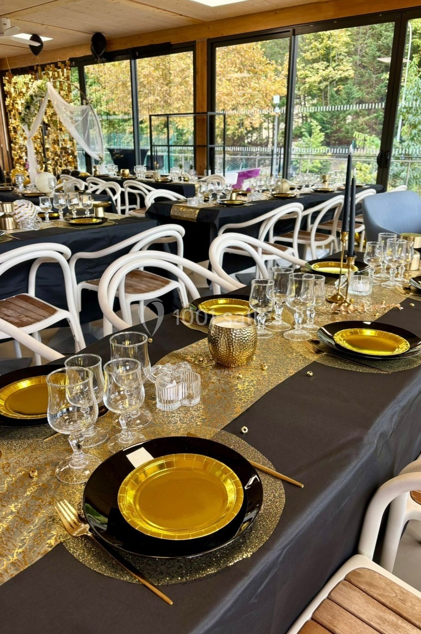 Salle décorée pour un événement avec tables dressées en noir et or, vaisselle élégante et grandes baies vitrées donnant sur…