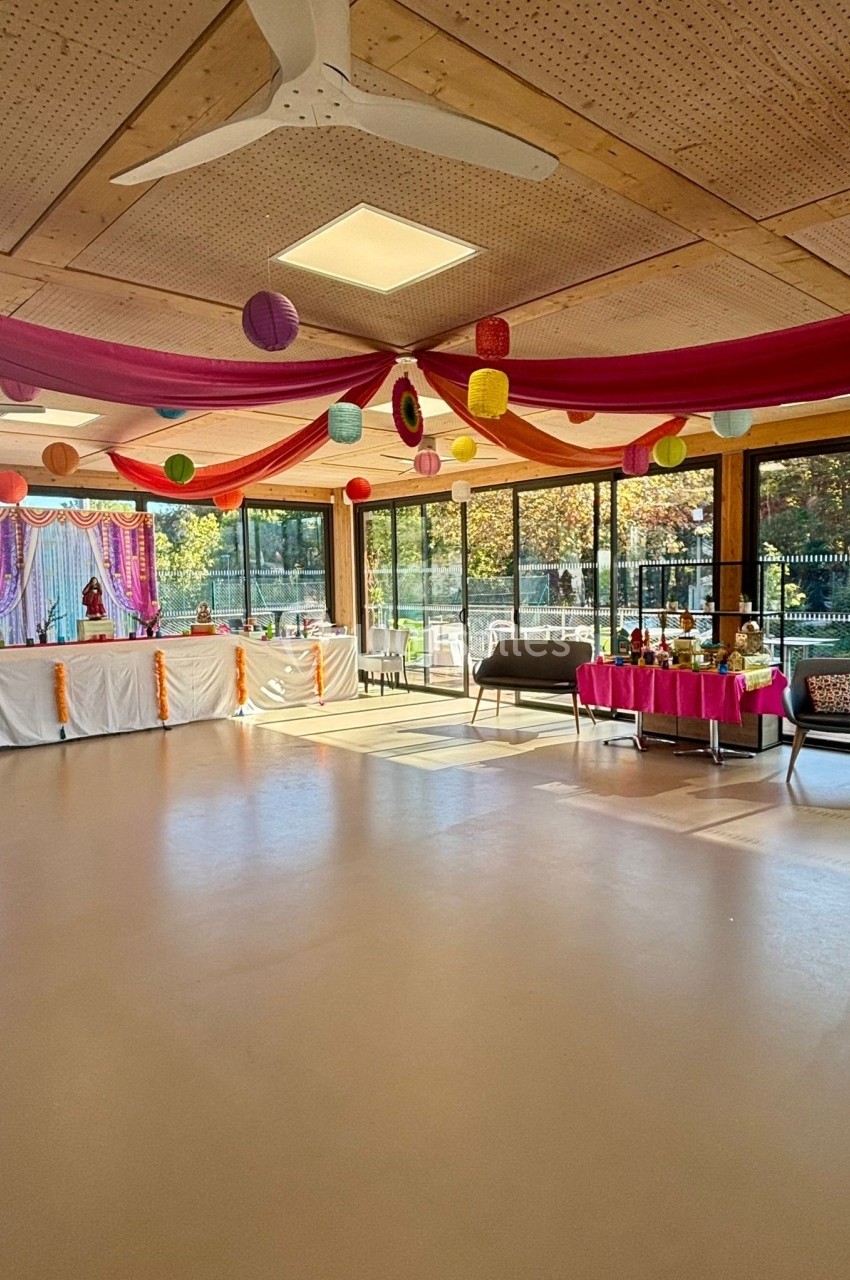 Salle décorée avec des guirlandes colorées et des lanternes, tables dressées pour un événement festif près de grandes baies…
