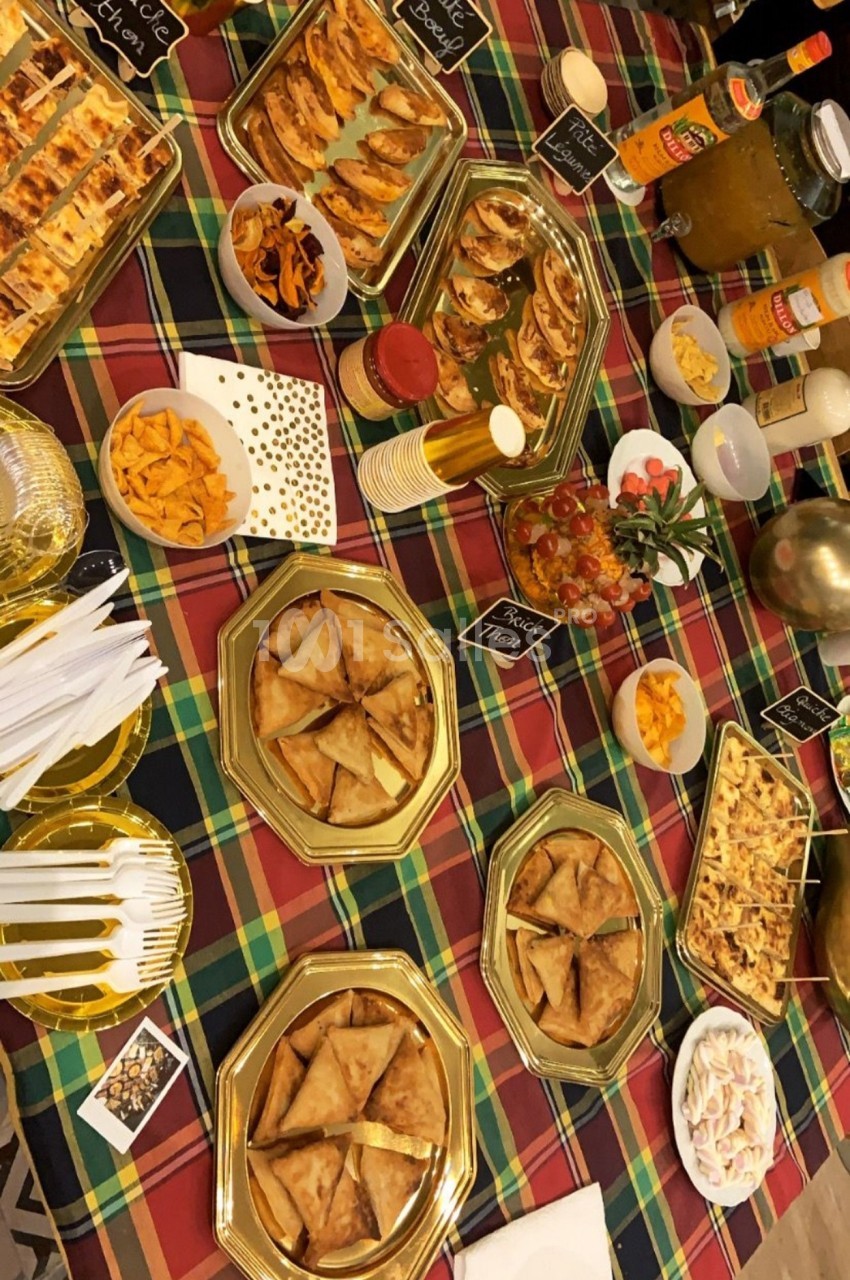 Table garnie de plats variés, notamment des feuilletés, des samoussas et des boissons, sur une nappe à carreaux colorée.