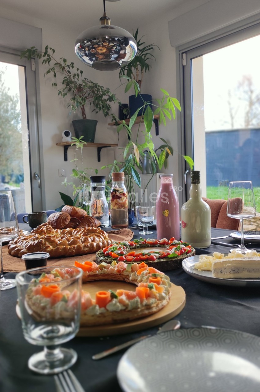 Table dressée avec des plats variés, incluant tartes salées, viennoiseries, boissons et fromages, dans une pièce lumineuse.