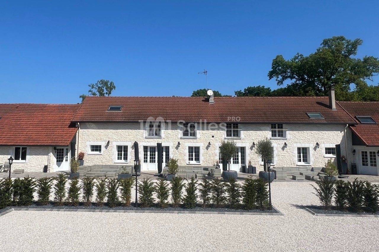 Façade d'une grande maison en pierre avec toit en tuiles rouges, entourée d'une cour gravillonnée et de haies.