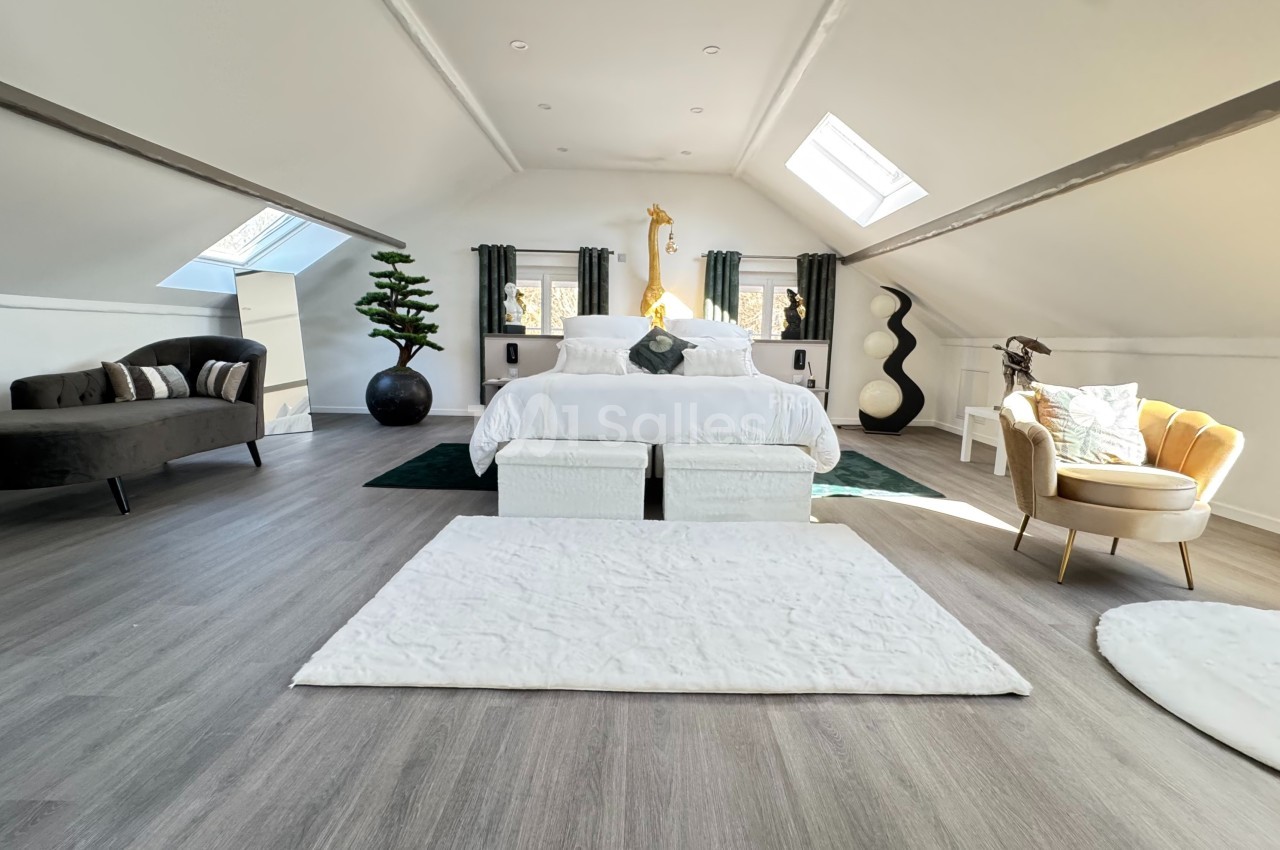 Chambre mansardée lumineuse avec lit central, fauteuils, canapé, décoration moderne et puits de lumière.