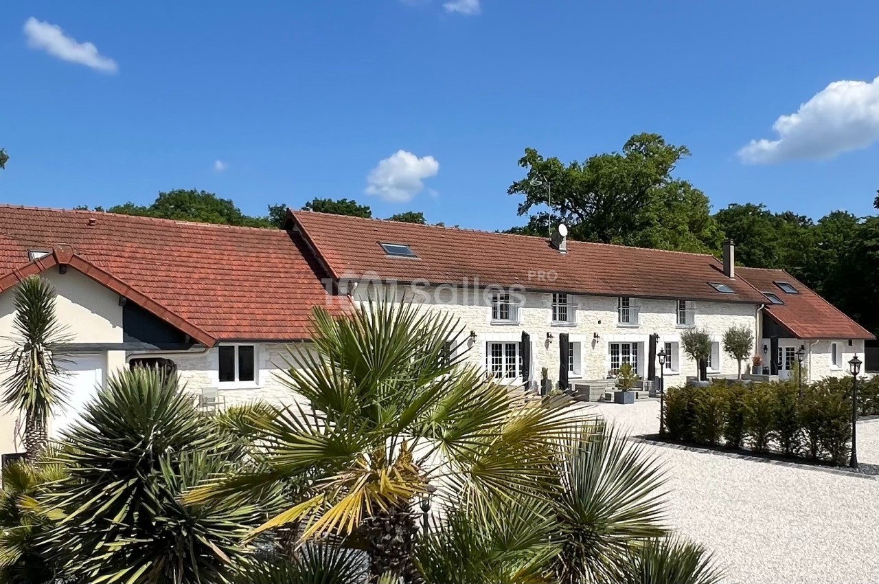 Grande maison en pierre avec toit en tuiles rouges, entourée de végétation et d'une cour gravillonnée sous un ciel bleu.