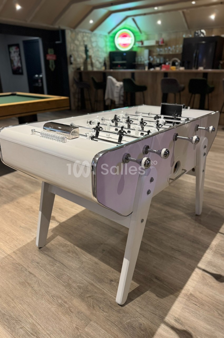 Table de baby-foot blanche dans une salle de loisirs avec un bar et une table de billard en arrière-plan.