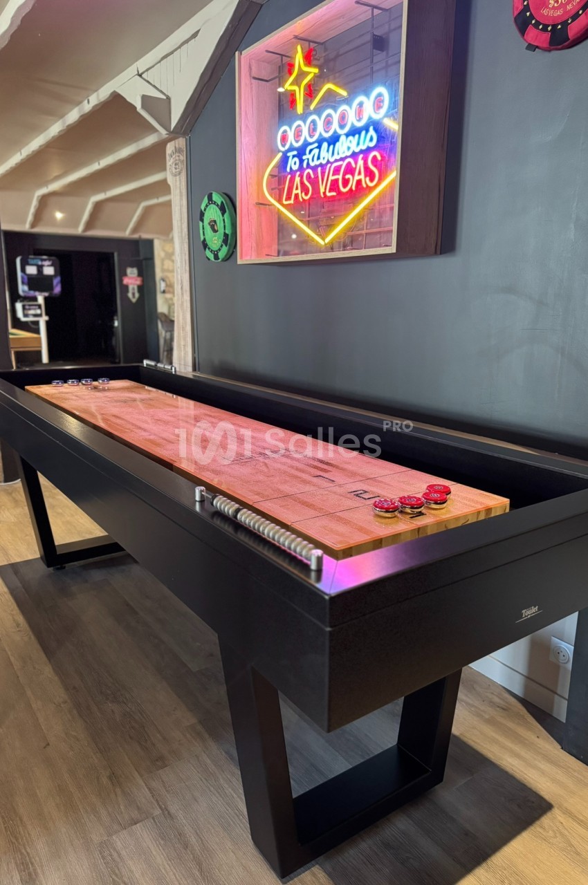 Table de shuffleboard en bois avec des pions, située dans une pièce décorée d'une enseigne lumineuse ’Las Vegas’.