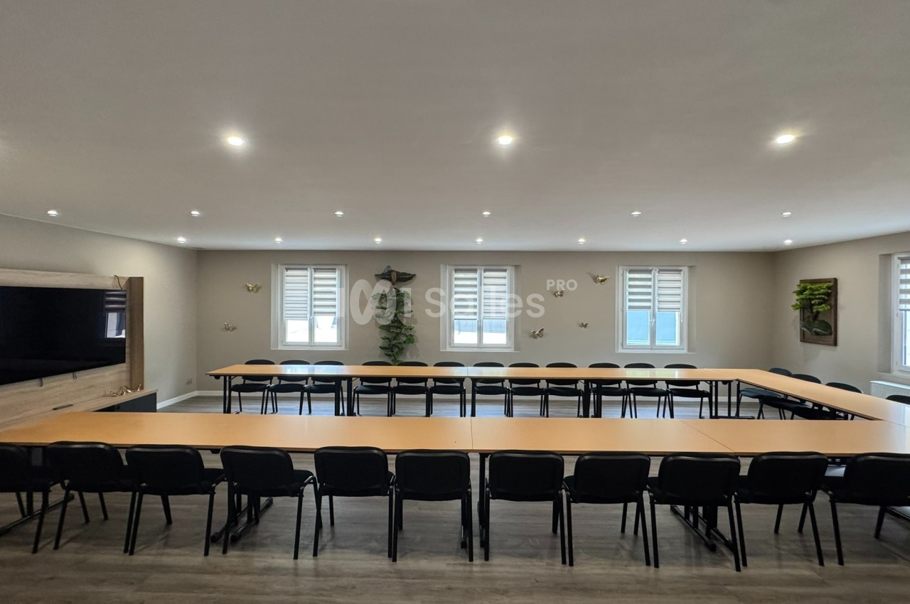 Salle de réunion lumineuse avec tables disposées en U, chaises noires, écran mural et décorations végétales.