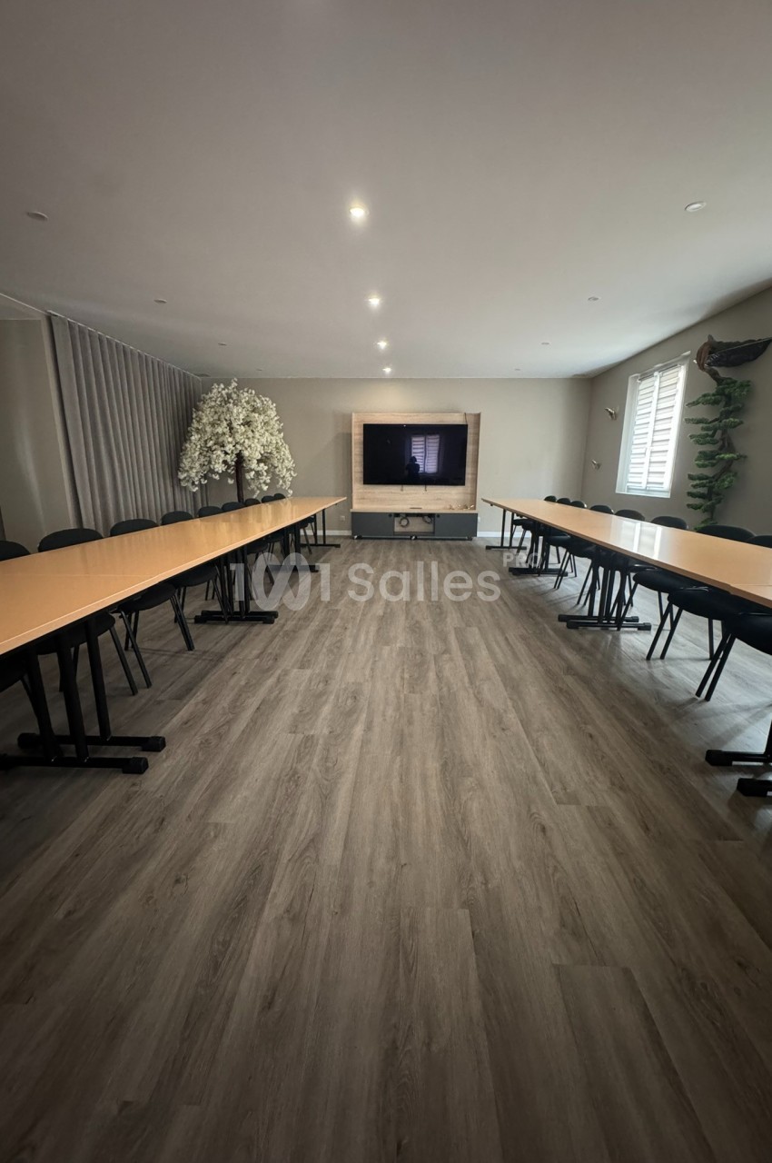 Salle de réunion lumineuse avec deux longues tables en U, écran mural et sol en parquet clair.
