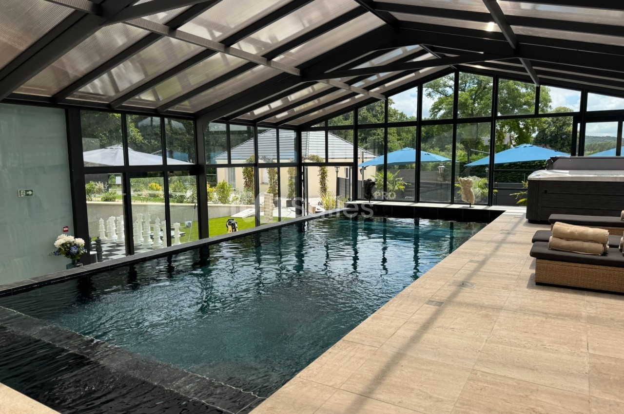 Piscine intérieure avec toit en verre, entourée de baies vitrées donnant sur un jardin avec parasols bleus.