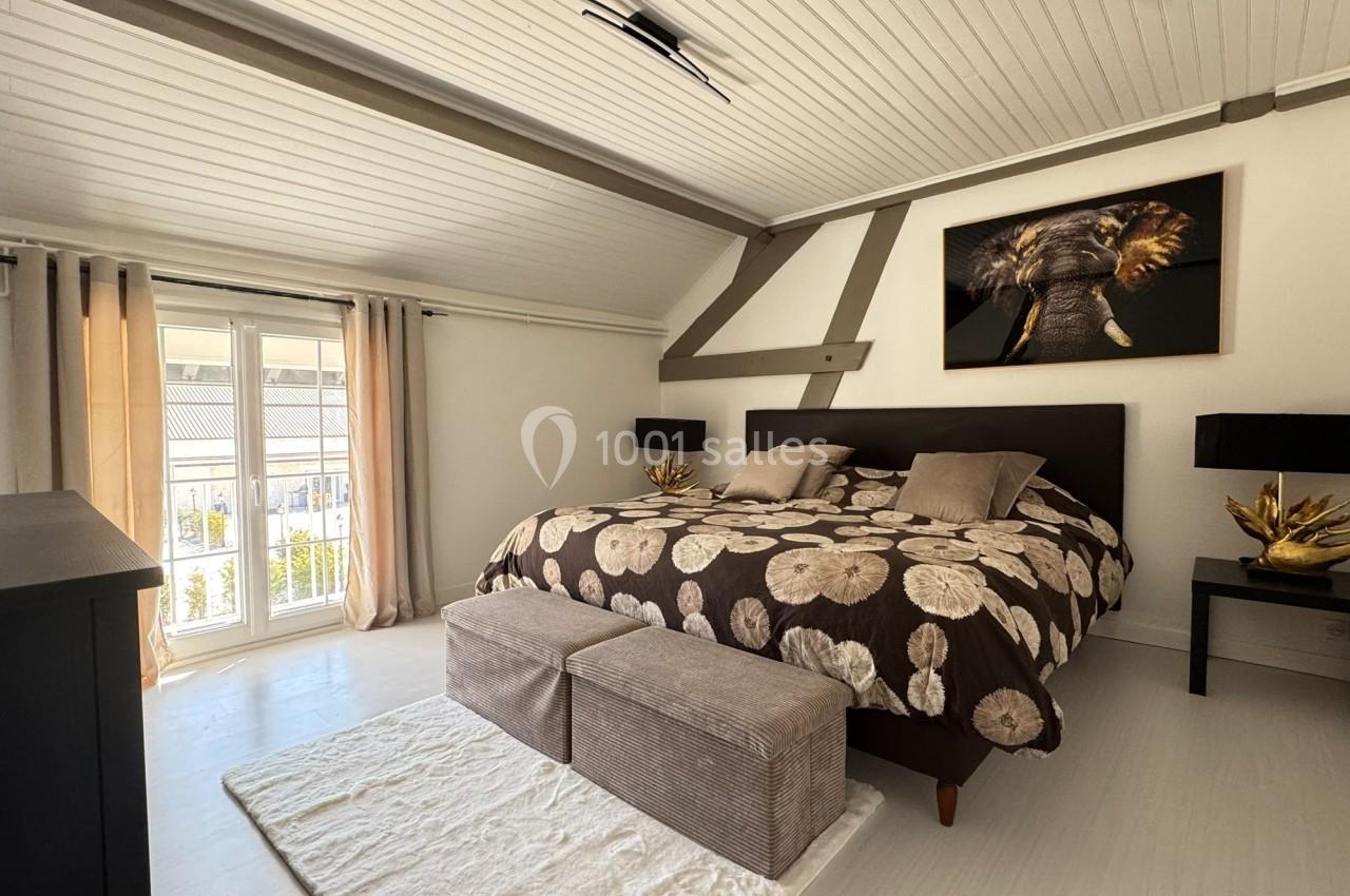 Chambre lumineuse avec lit double, couvre-lit à motifs floraux, deux poufs, tableau d'éléphant et fenêtres donnant sur l…