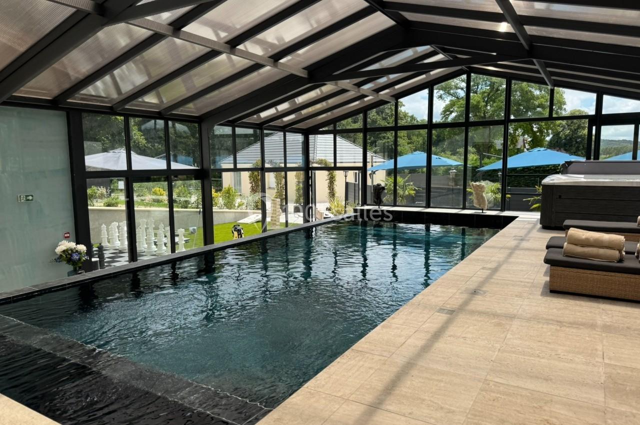 Piscine intérieure avec toit en verre, entourée de baies vitrées donnant sur un jardin avec parasols bleus.