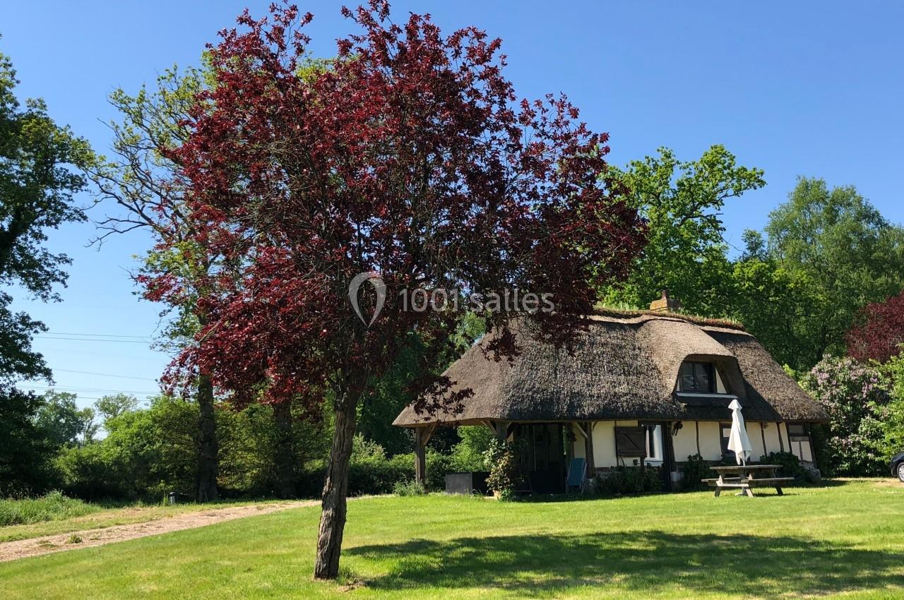 Maison à toit de chaume entourée de verdure, avec un arbre aux feuilles rouges au premier plan.