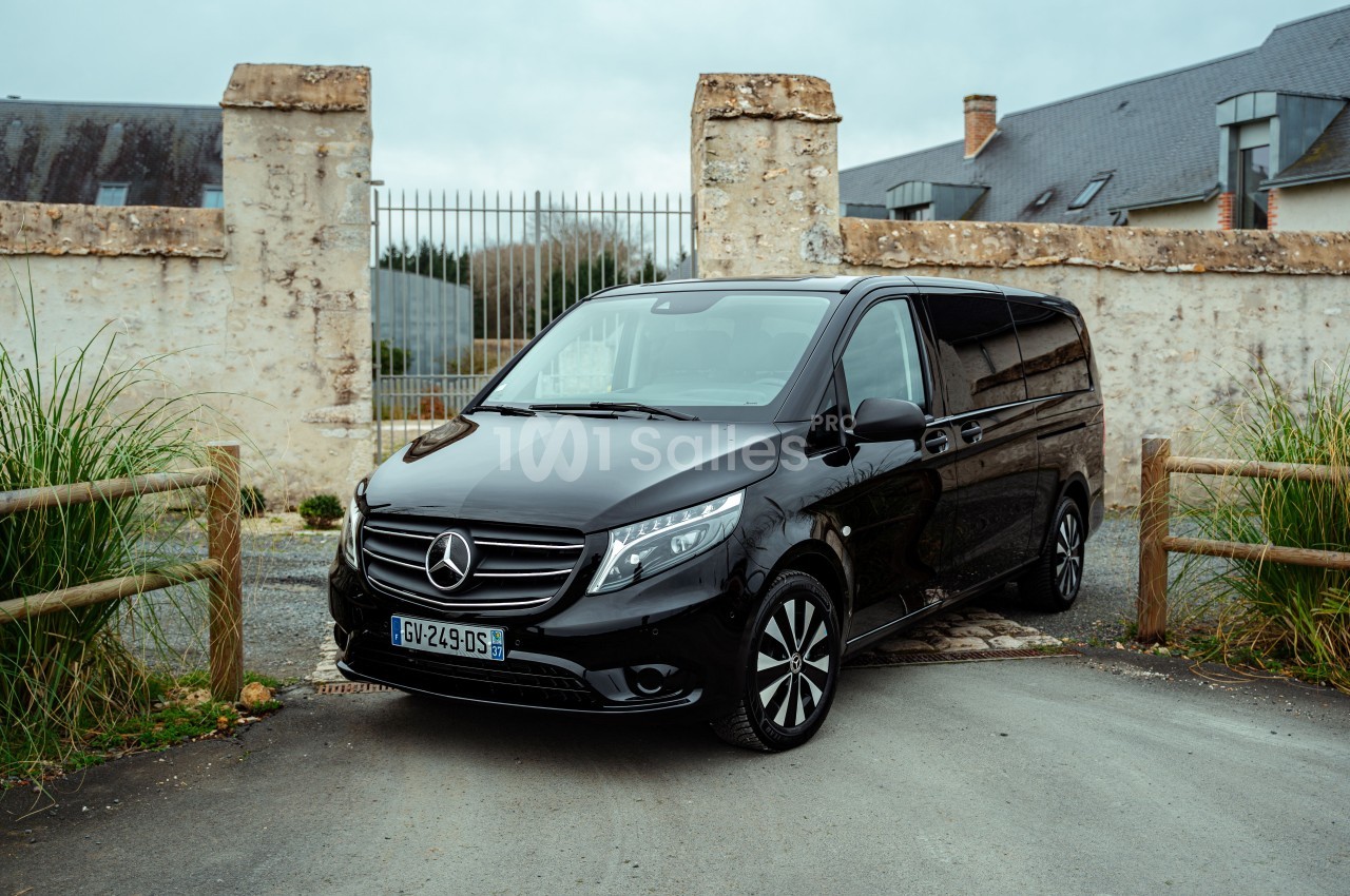 Un van noir Mercedes garé devant un portail en métal, entouré de murs en pierre et de végétation.