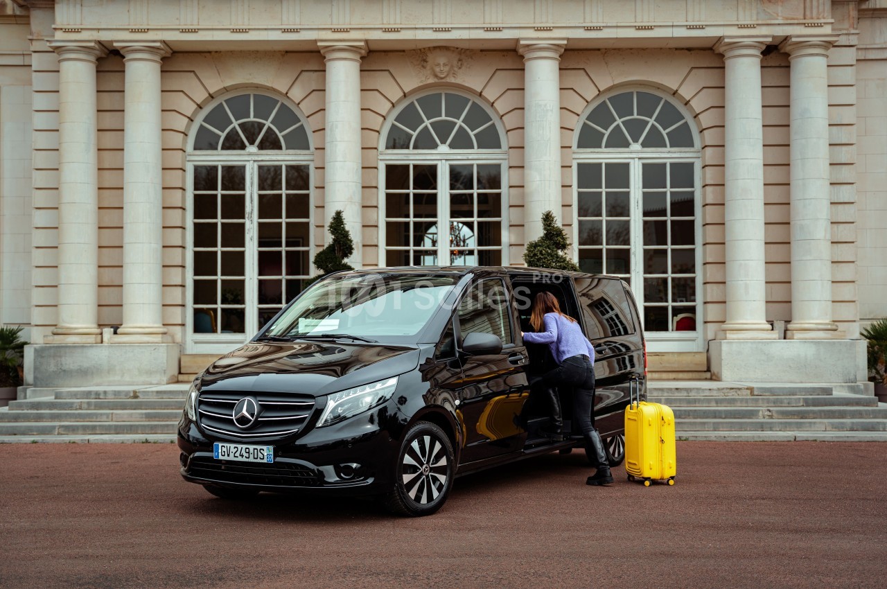 Une femme range une valise jaune dans un van noir stationné devant un bâtiment à colonnes blanches.