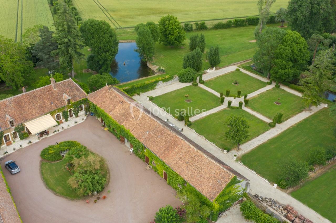 Vue aérienne d'un domaine avec bâtiments en pierre, jardins aménagés, allées gravillonnées et un étang entouré de verdure.